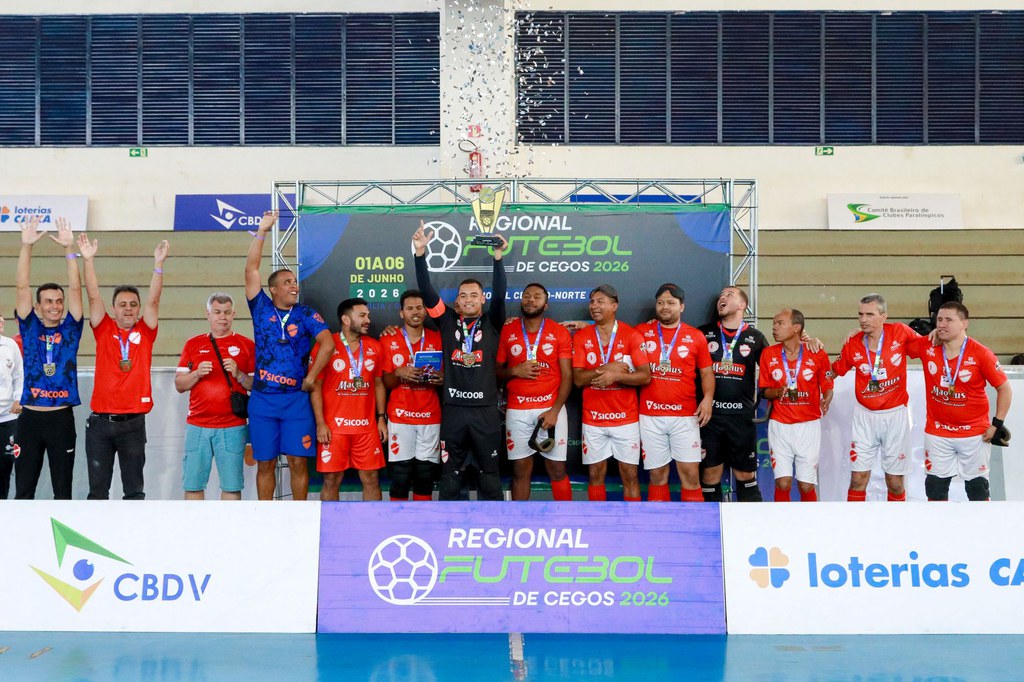 Na cerimônia de premiação do Regional Centro-Norte de Futebol de Cegos 2026, atletas e integrantes da comissão técnica do Vila Nova/Acelgo-GO posam juntos no pódio após a conquista do tricampeonato. Ao centro, um dos jogadores ergue o troféu enquanto papéis metalizados caem do alto. Os atletas vestem uniformes vermelhos com detalhes brancos e exibem as medalhas conquistadas. Ao fundo, o painel oficial da competição destaca a identidade visual do torneio, realizado em Brasília.